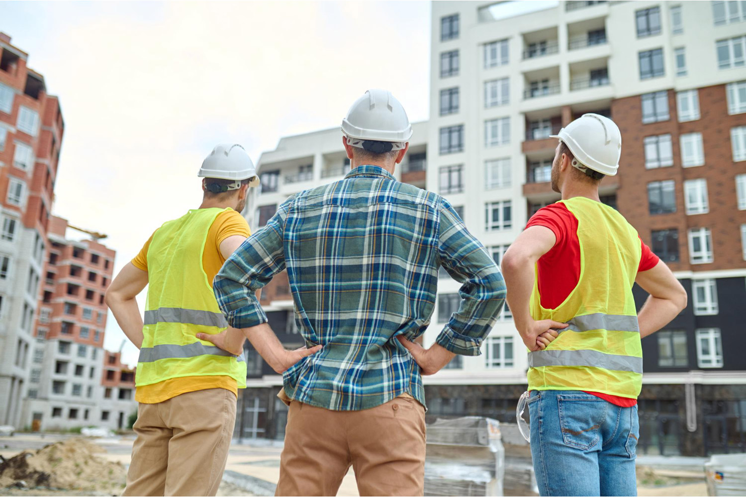 verduurzaming-op-maat-bouwplaats-vastgoed-procalor Drie bouwprofessionals met helmen kijken naar een modern appartementencomplex tijdens een verduurzamingsproject op maat.