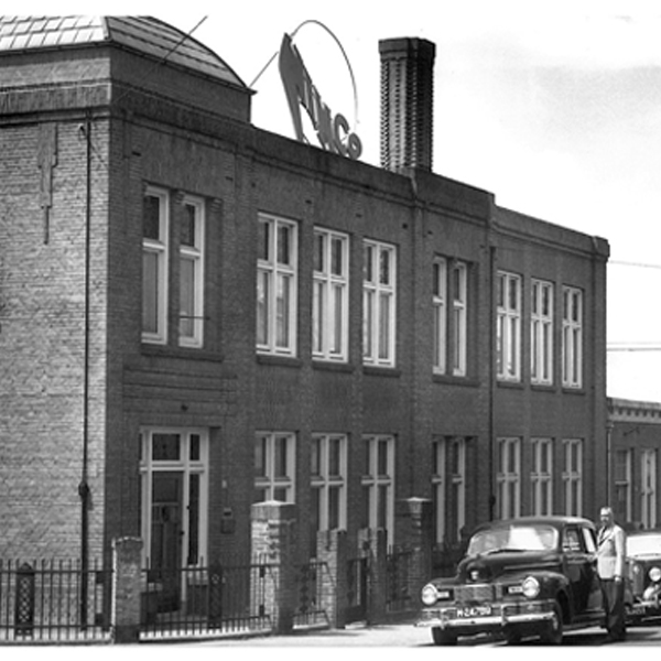 Zwart-witfoto van de Nimco-fabriek in Nijmegen met het herkenbare Nimco-logo op het dak en een geparkeerde auto op de voorgrond.
